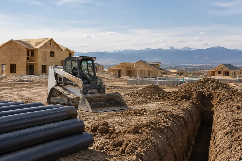 Skid steer and stacked pipe beside a fresh trench near homes under construction